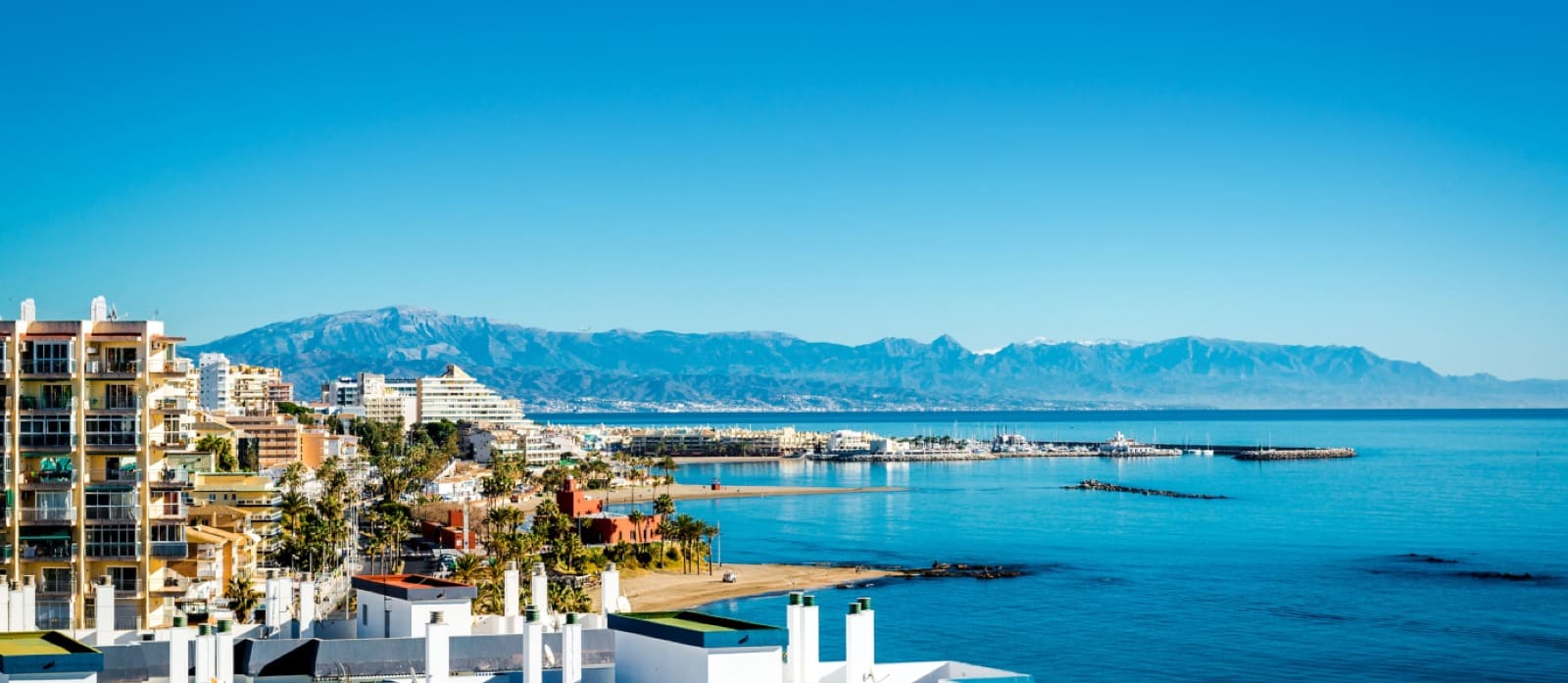 Apartment blocks, a sandy beach and a marina along a Costa del Sol bay, with mountains behind
