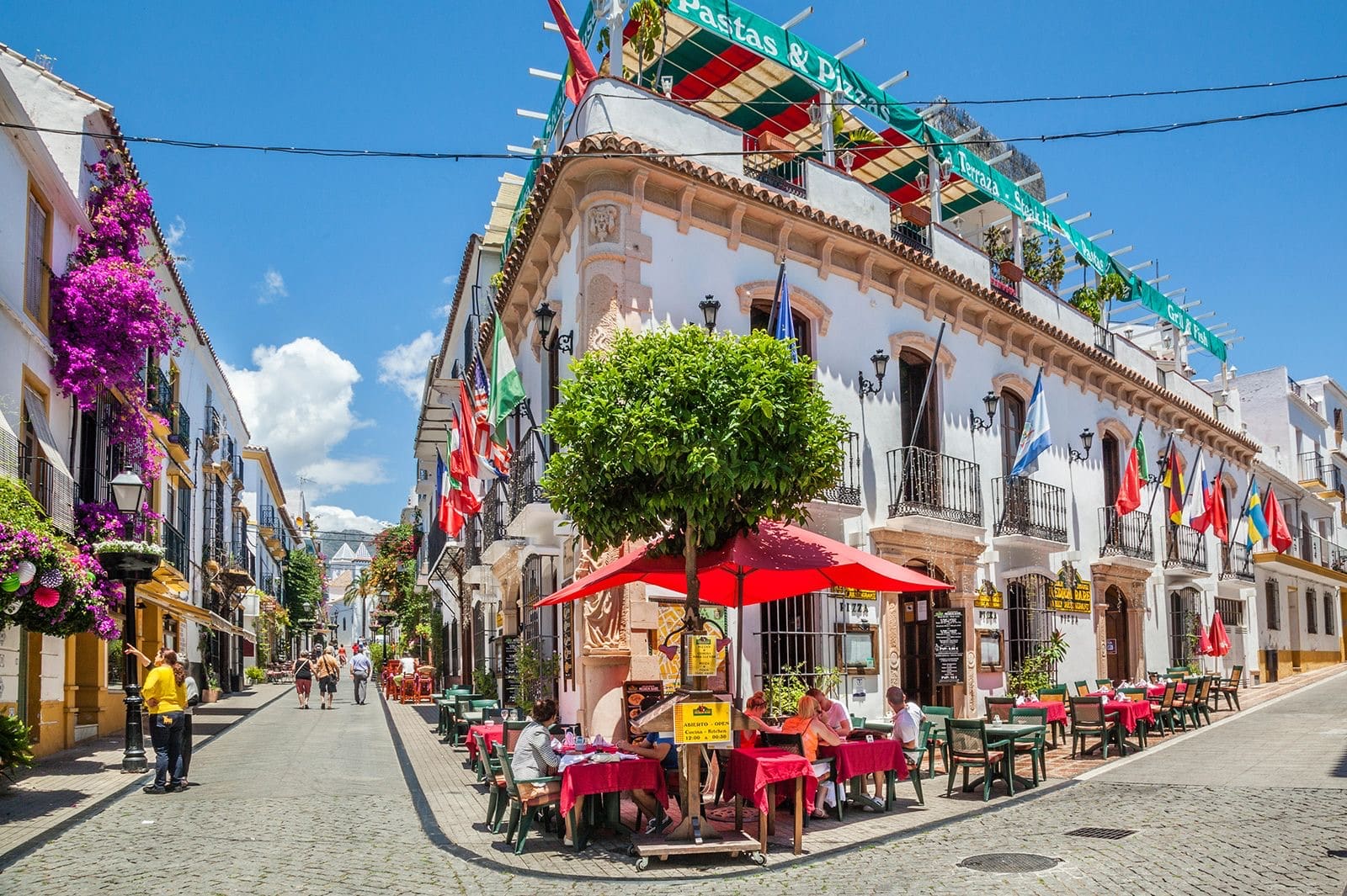 A sunlit Costa del Sol old-town street with flowers, flags and outdoor café tables