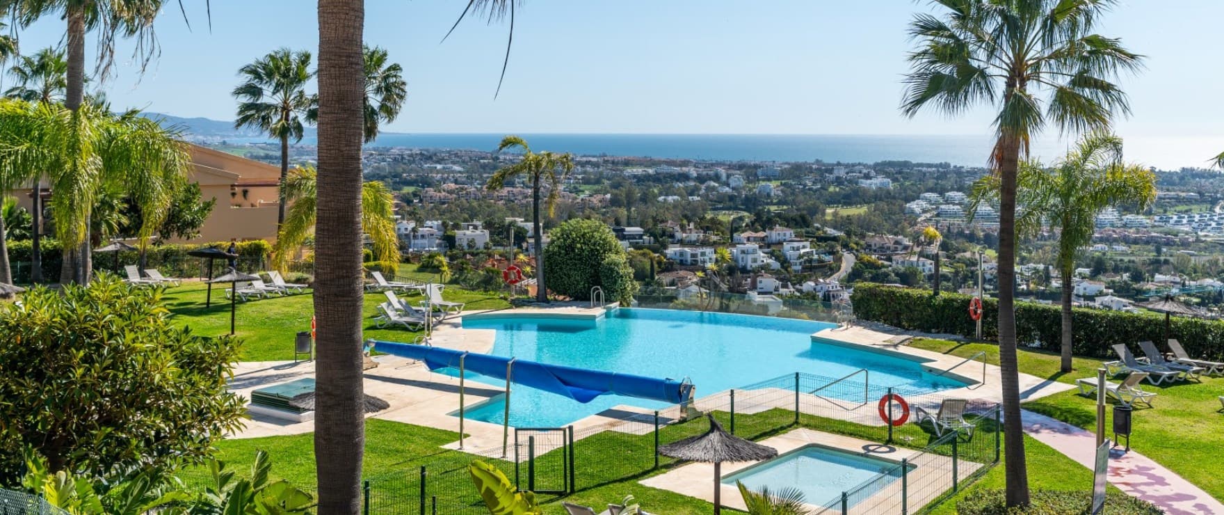 One of the estate’s pools, with a children’s pool beside it, palms around the lawn, and the coast and the sea beyond. Altura 160, Señorío de Cifuentes, Benahavís.