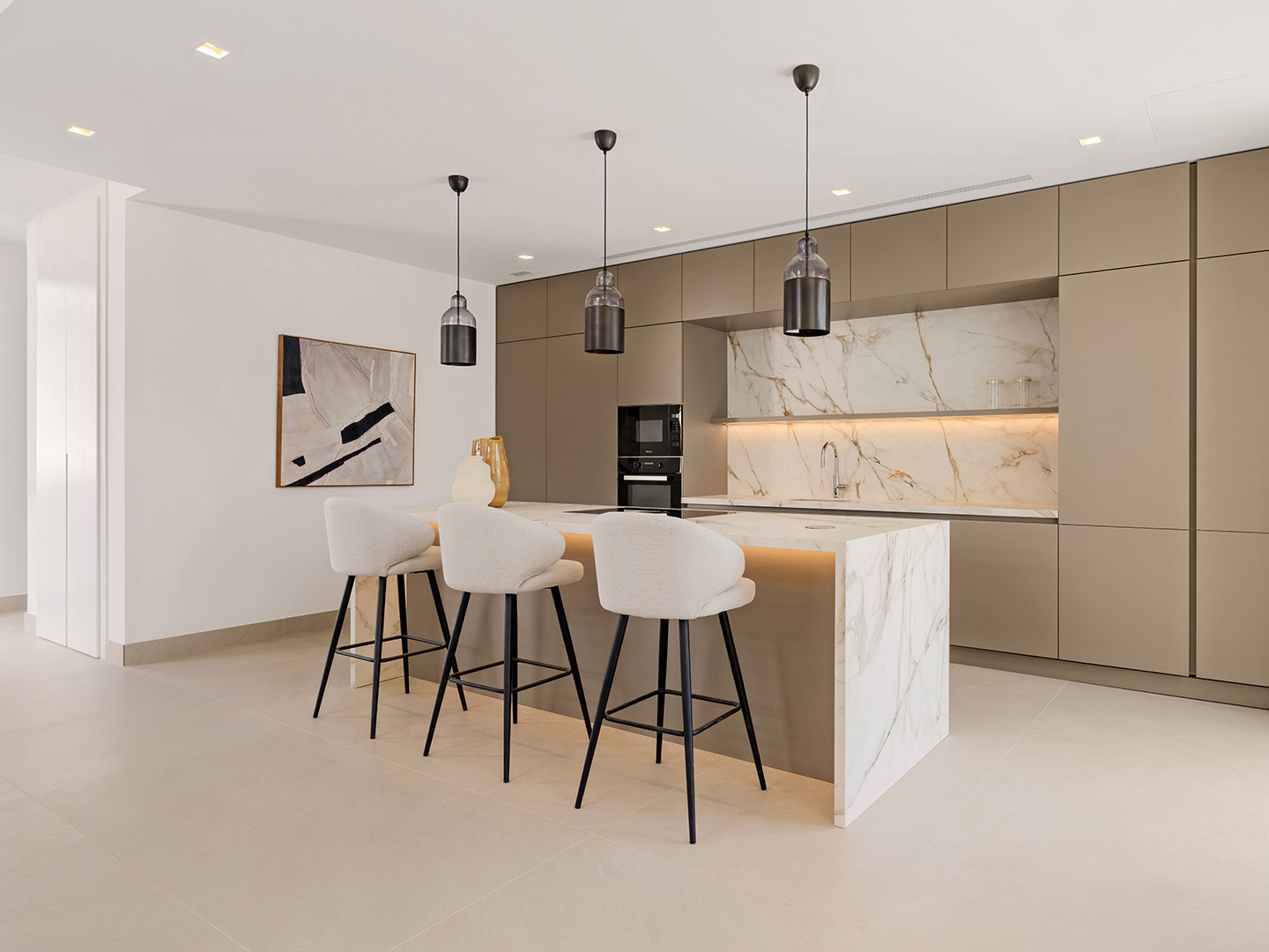 The kitchen of the show villa, with an island and splashback in white stone veined in gold and matt units in a warm grey. Aurea Villas, Estepona Golf, Estepona.
