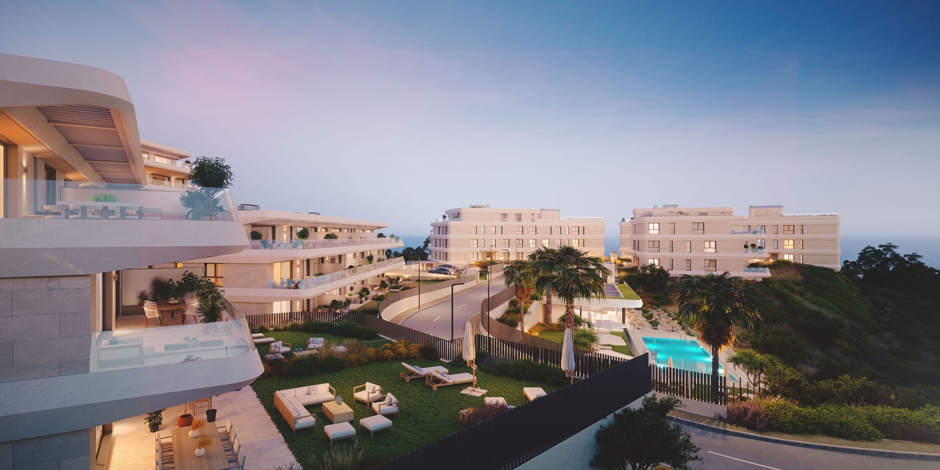Dusk over a lawn with outdoor sofas, with the pool beyond and lit terraces on the building to the left. Australy Aures II, La Resina, Estepona.