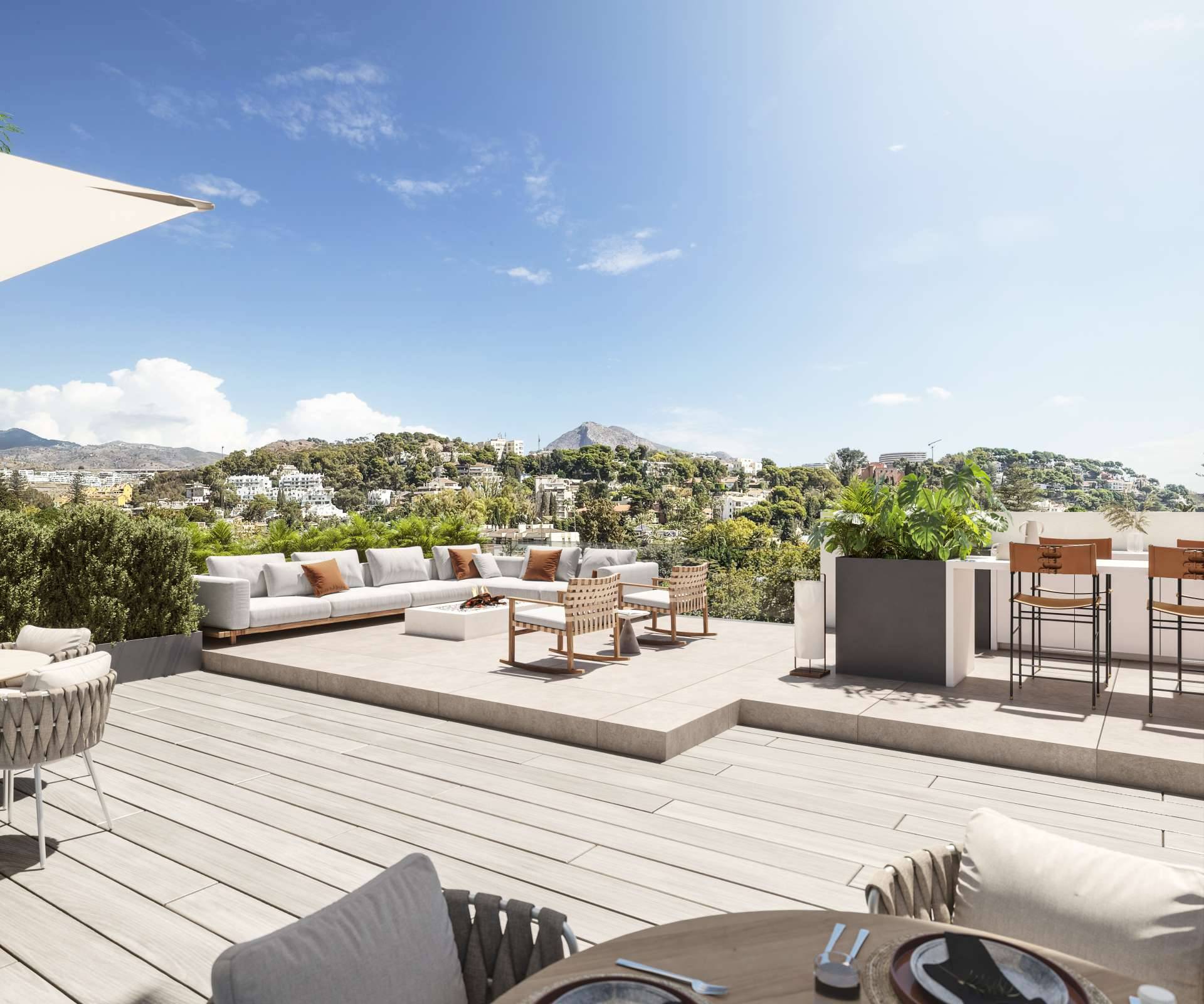 A roof terrace with a long sofa, two rocking chairs and a bar with high stools, with wooded hills behind. Be Grand El Limonar, El Limonar, Málaga.