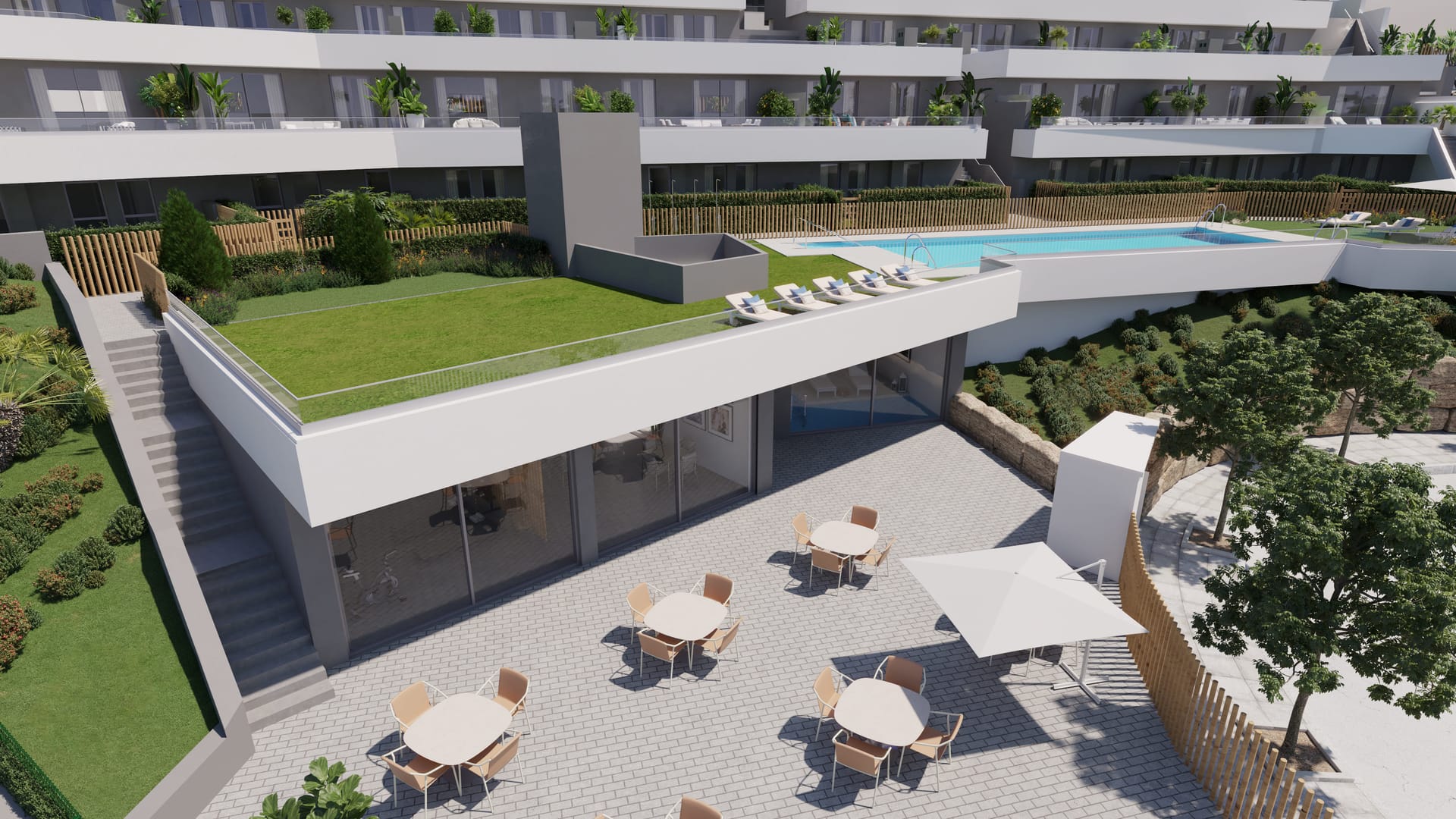 Tables and chairs on a paved terrace outside a glass-fronted communal building with a lawn on its roof, and the pool on the level above. Blue Marine, La Chullera, Manilva.
