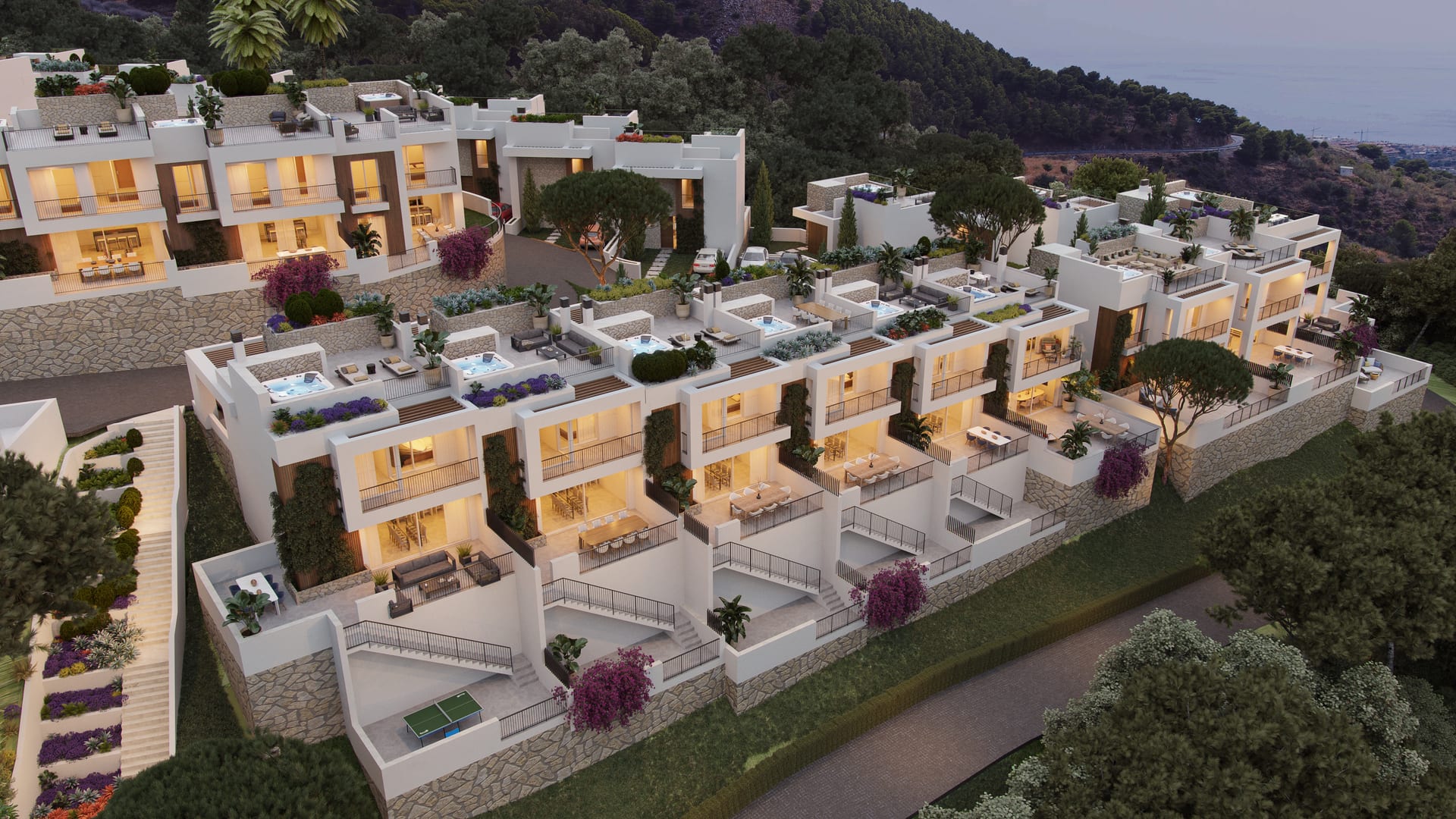 An evening view of a group of villas, with their living floors and roof terraces lit and the sea on the horizon. Buenavista Views, Buenavista, Mijas.