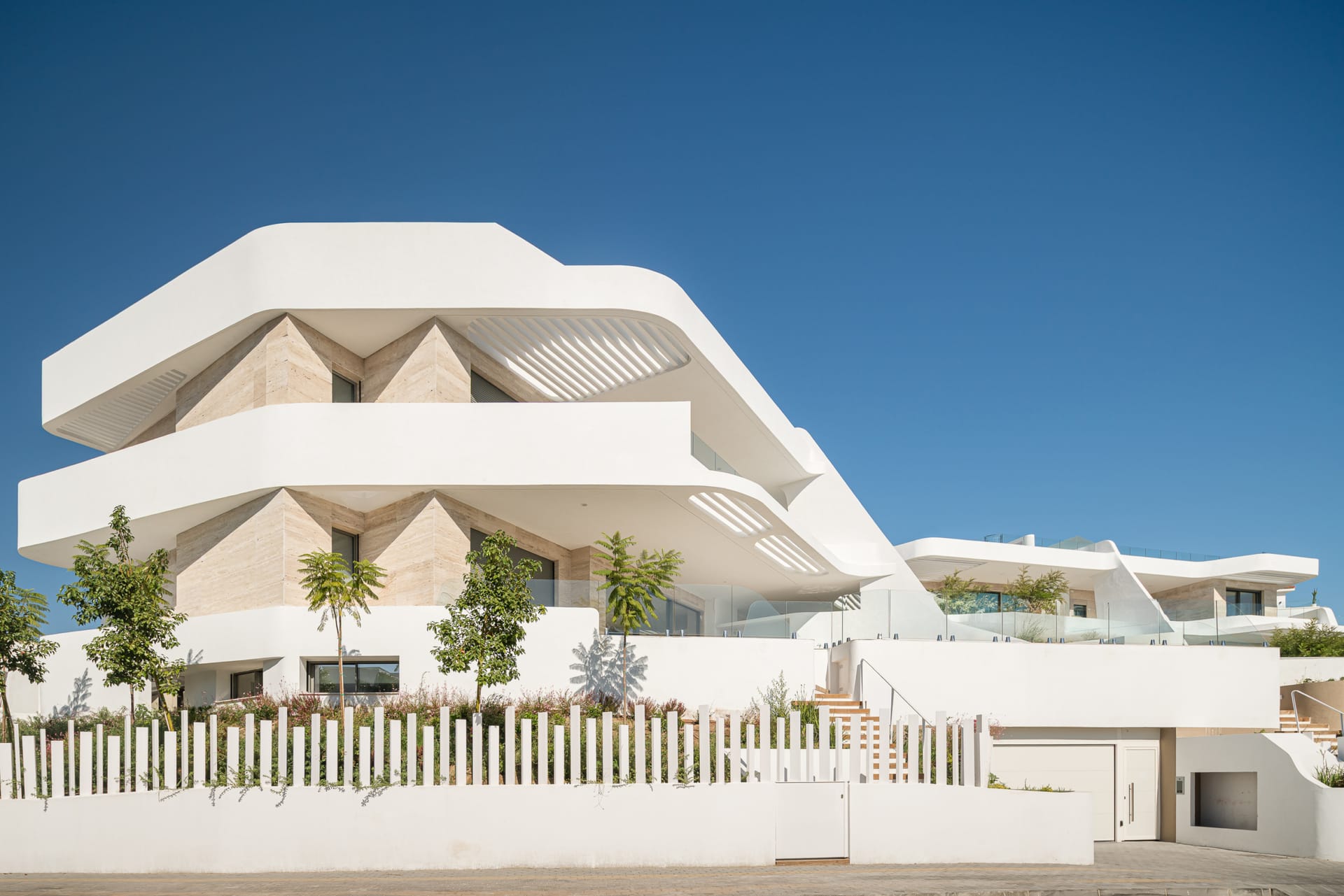 The front of the development from the street, with its curved white terraces, travertine walls and a white fence of upright posts. Golden Eight, Cabopino, Marbella.