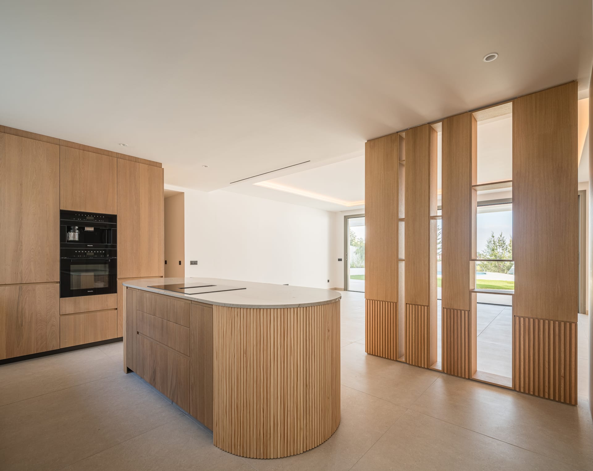A kitchen in one of the homes, with oak units, the oven and microwave set into the wall, a rounded island holding the induction hob, and a slatted oak screen with the garden beyond. Golden Eight, Cabopino, Marbella.
