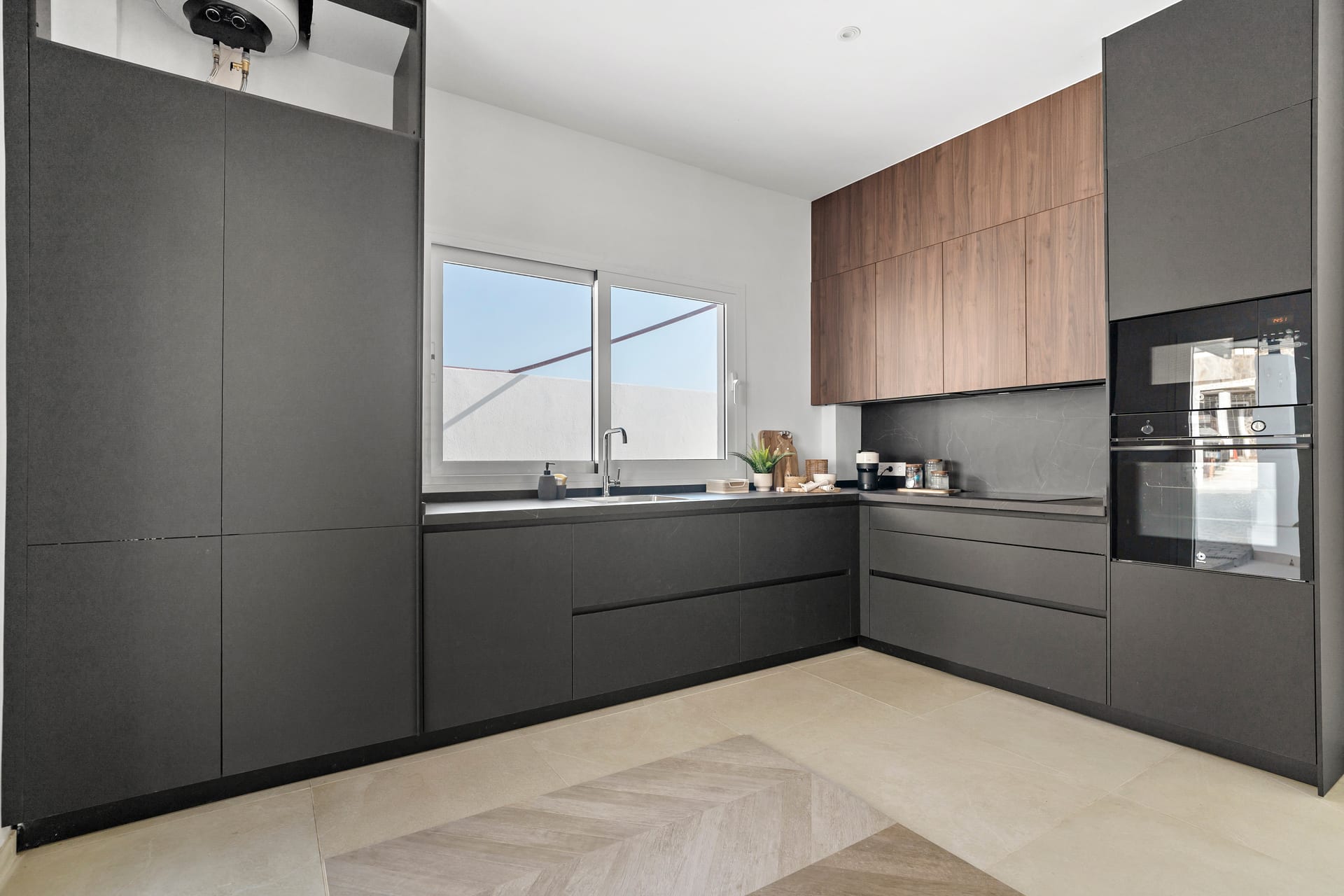 The show house kitchen, with dark grey units, a wood-finish wall cupboard and the oven and microwave set into a tall unit. Olimpo, Urbanización Olimpo, Mijas.