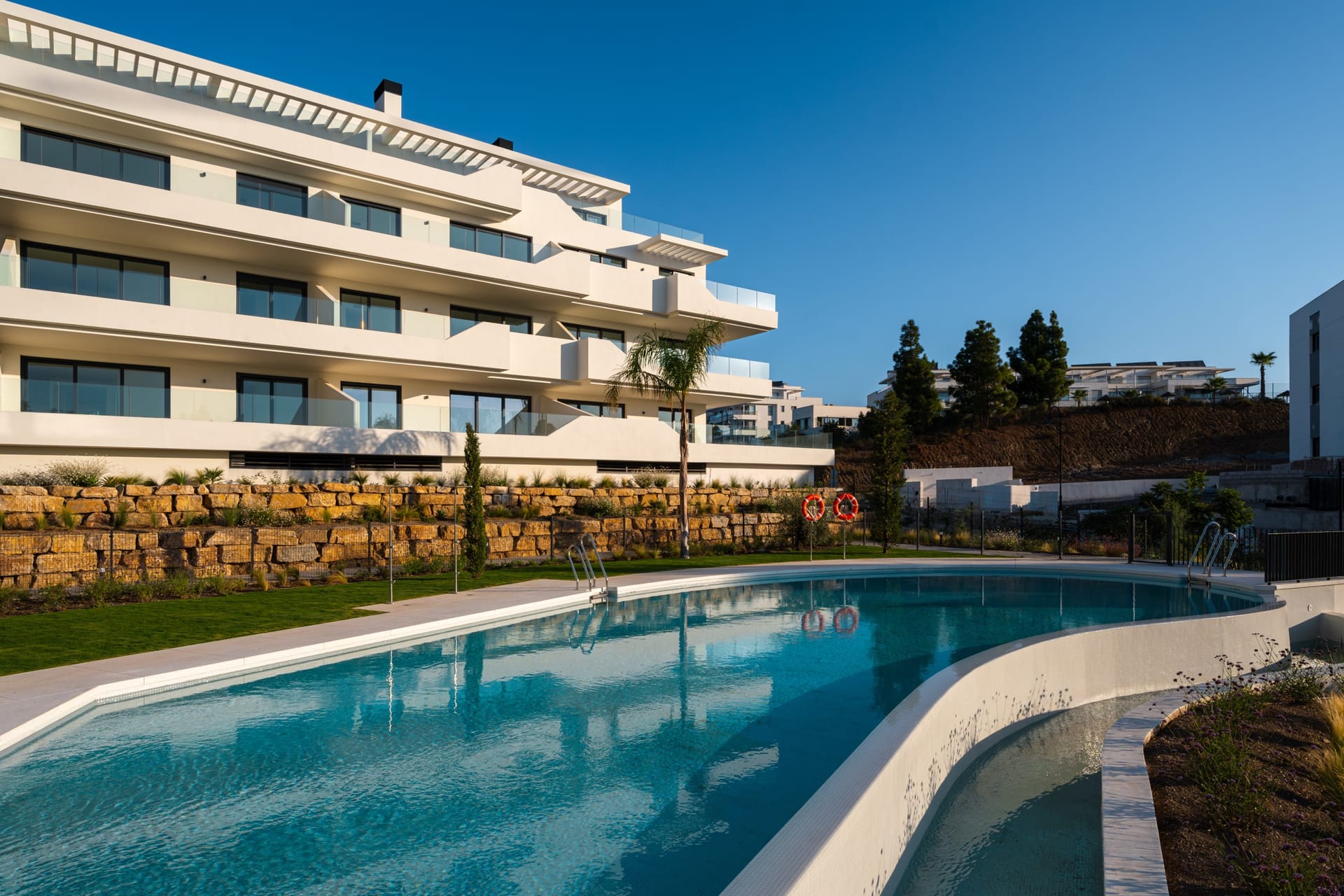 The main communal pool, curving along the foot of the buildings with lawn and planting behind. Rayos del Sol, El Chaparral, Mijas.