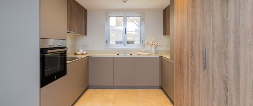 A U-shaped kitchen beneath a window, with grey-brown base units under a beige worktop and oak-coloured wall cupboards. Sunny Golf, Estepona Golf, Estepona.