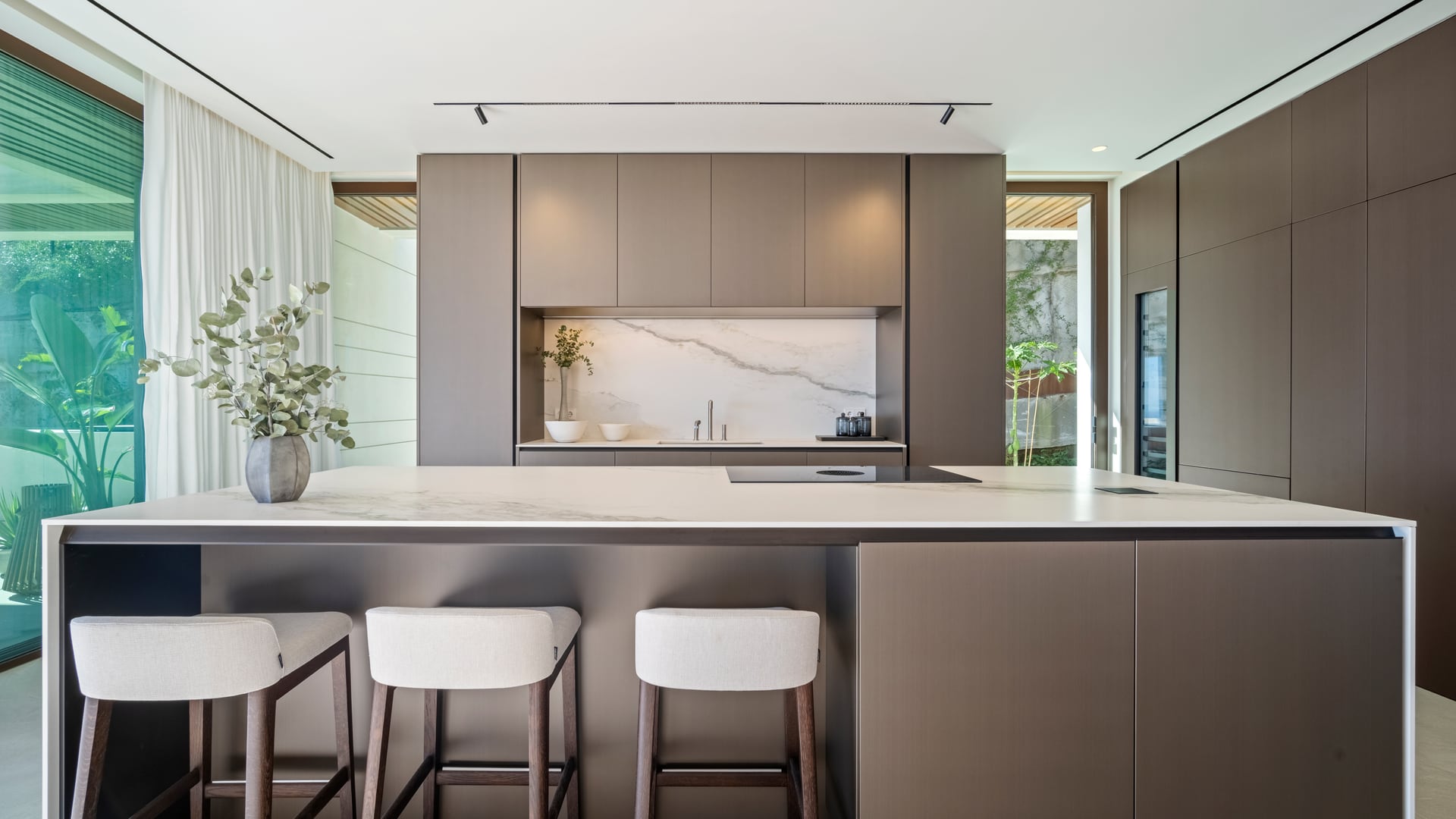 A villa kitchen, with taupe units from floor to ceiling, a marble-effect splashback, and a pale-topped island with three stools. The Sky Marbella, La Alborada, Benahavís.