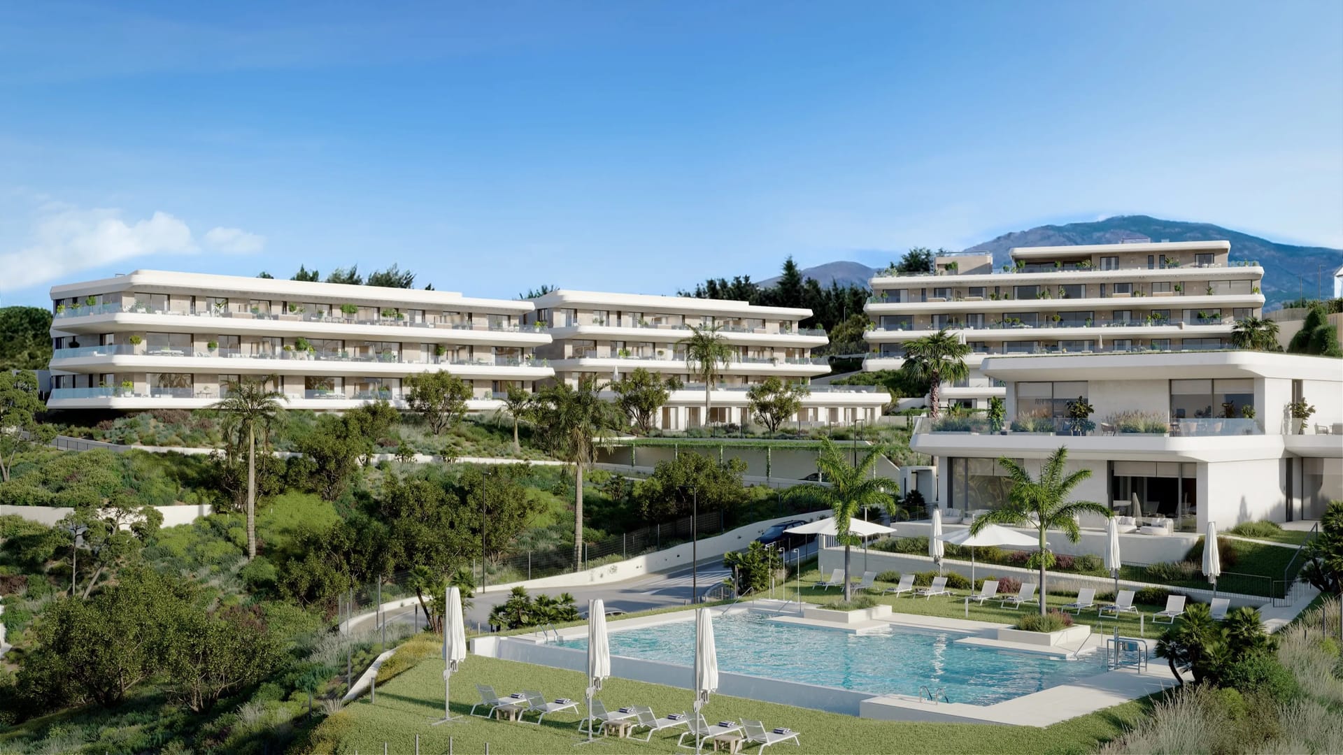 A pool set in a lawn with loungers and parasols, below long white buildings with mountains behind. Thera I, La Resina, Estepona.