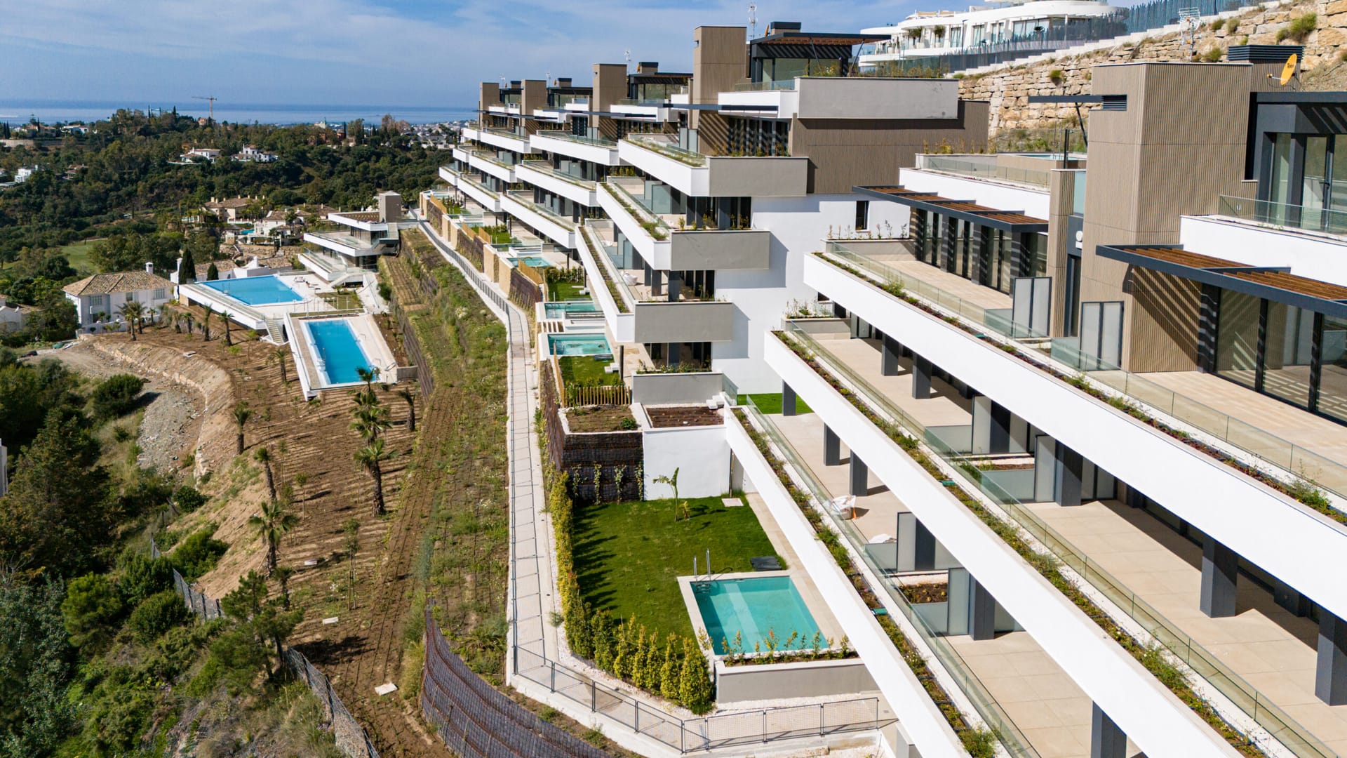 An aerial view of the buildings stepping down the hillside, with two of the pools and the coast in the distance. Tiara, La Quinta, Benahavís.