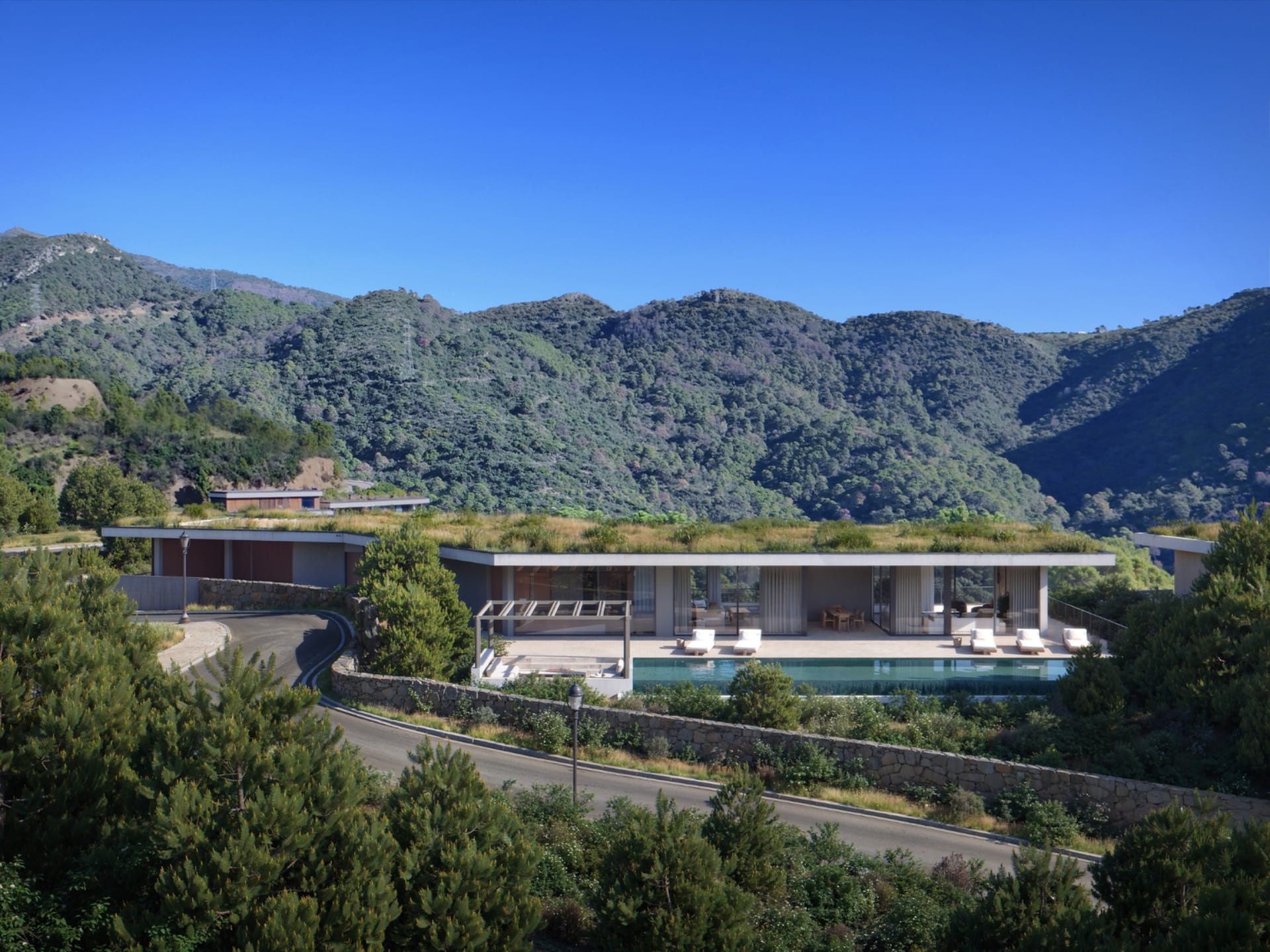 A villa set into the hillside below the mountains, with its pool and a road winding past. Vitae Villas, Monte Mayor, Benahavís.