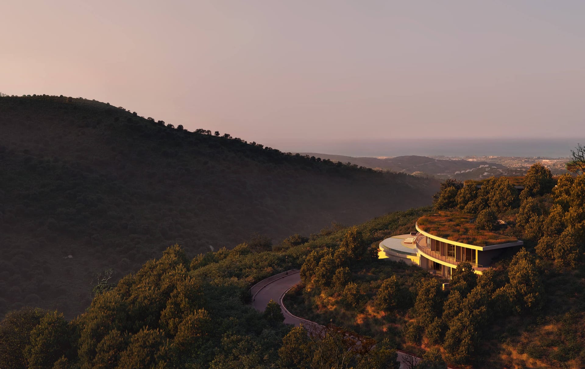 A villa lit up at dusk on the wooded hillside, with the coast and the sea in the distance. Vitae Villas, Monte Mayor, Benahavís.