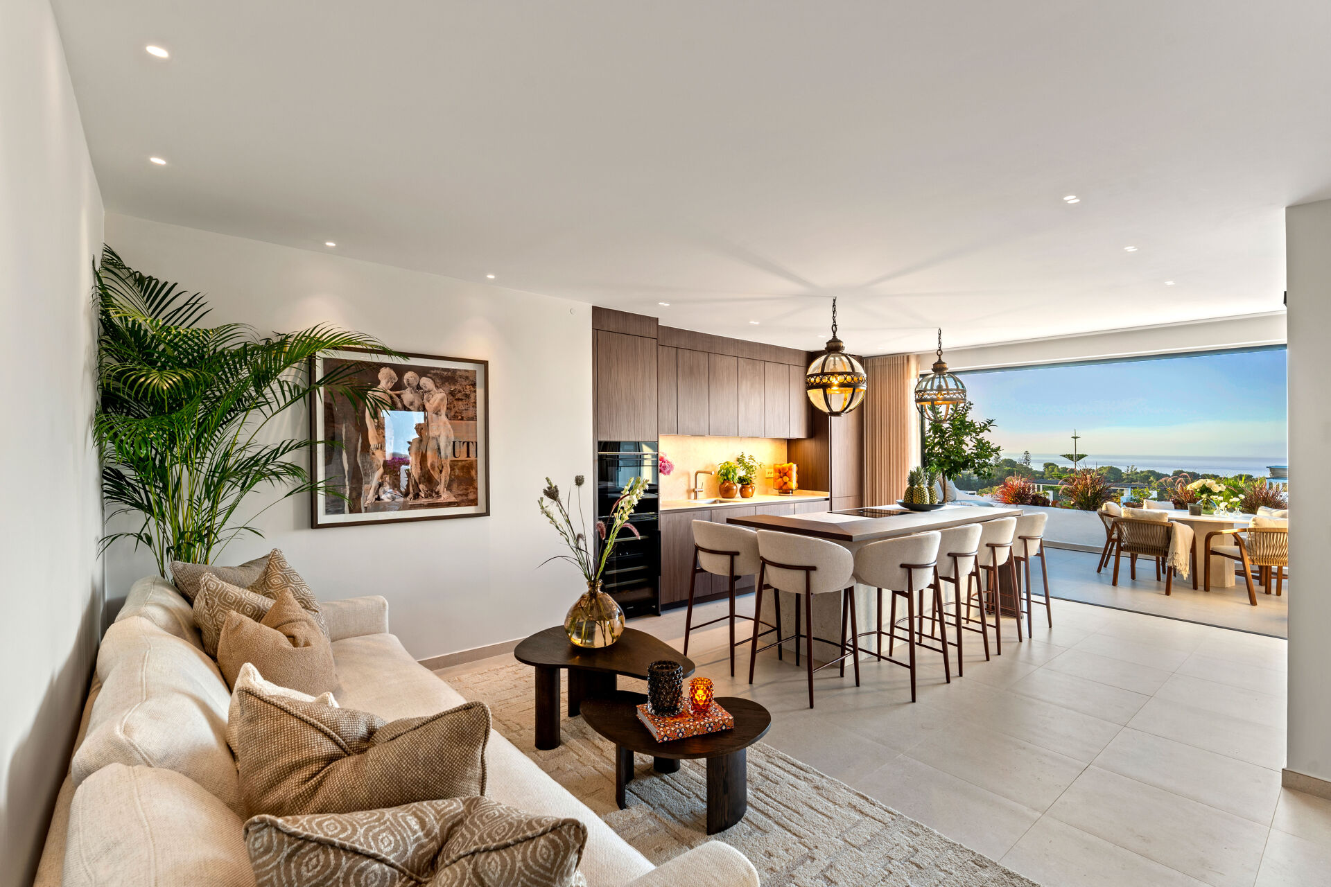 The living room, with a sofa in the foreground, the kitchen island with bar stools, and the terrace and the sea beyond.