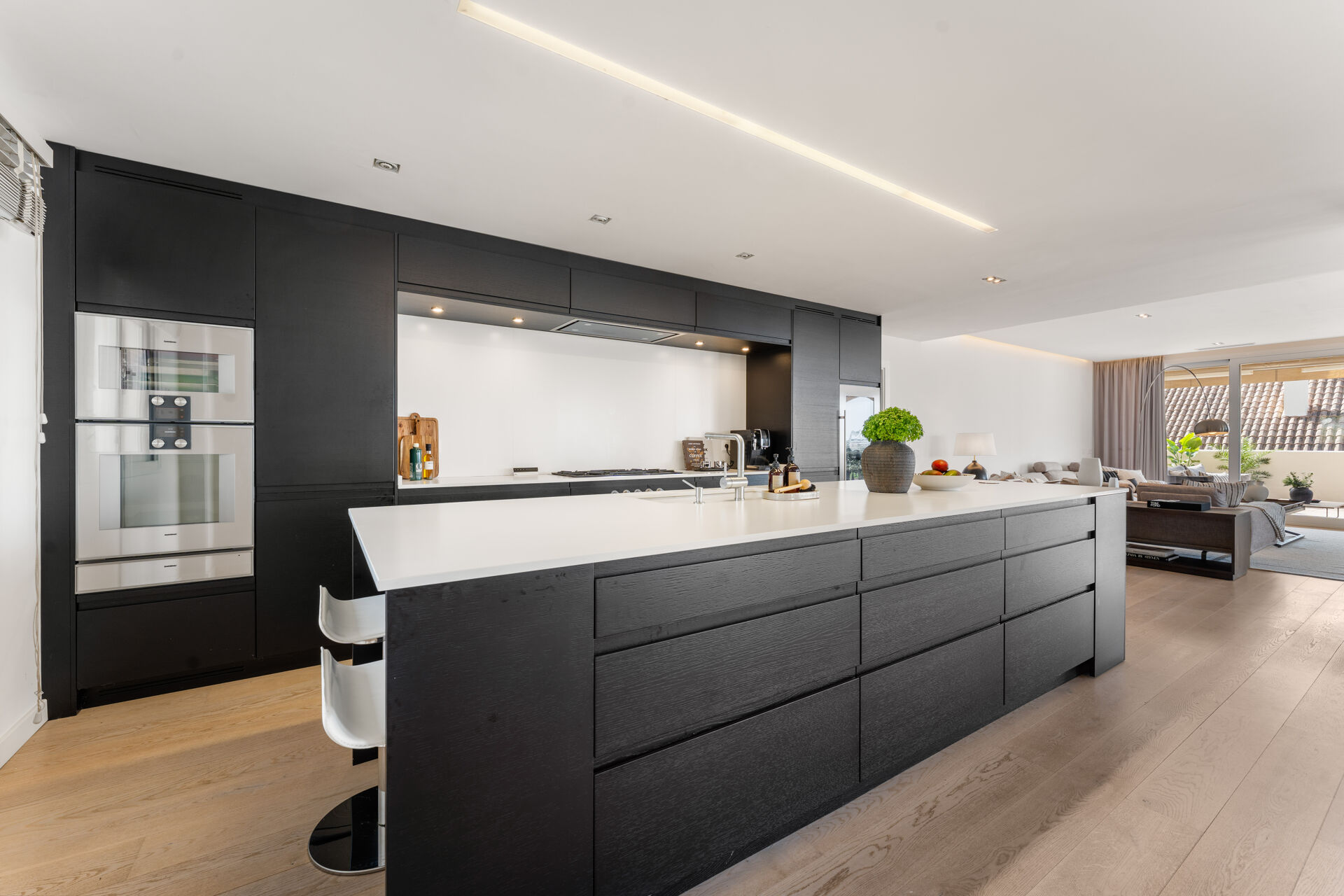 The kitchen, with black cabinets, two built-in ovens and a long white island, open to the living area.