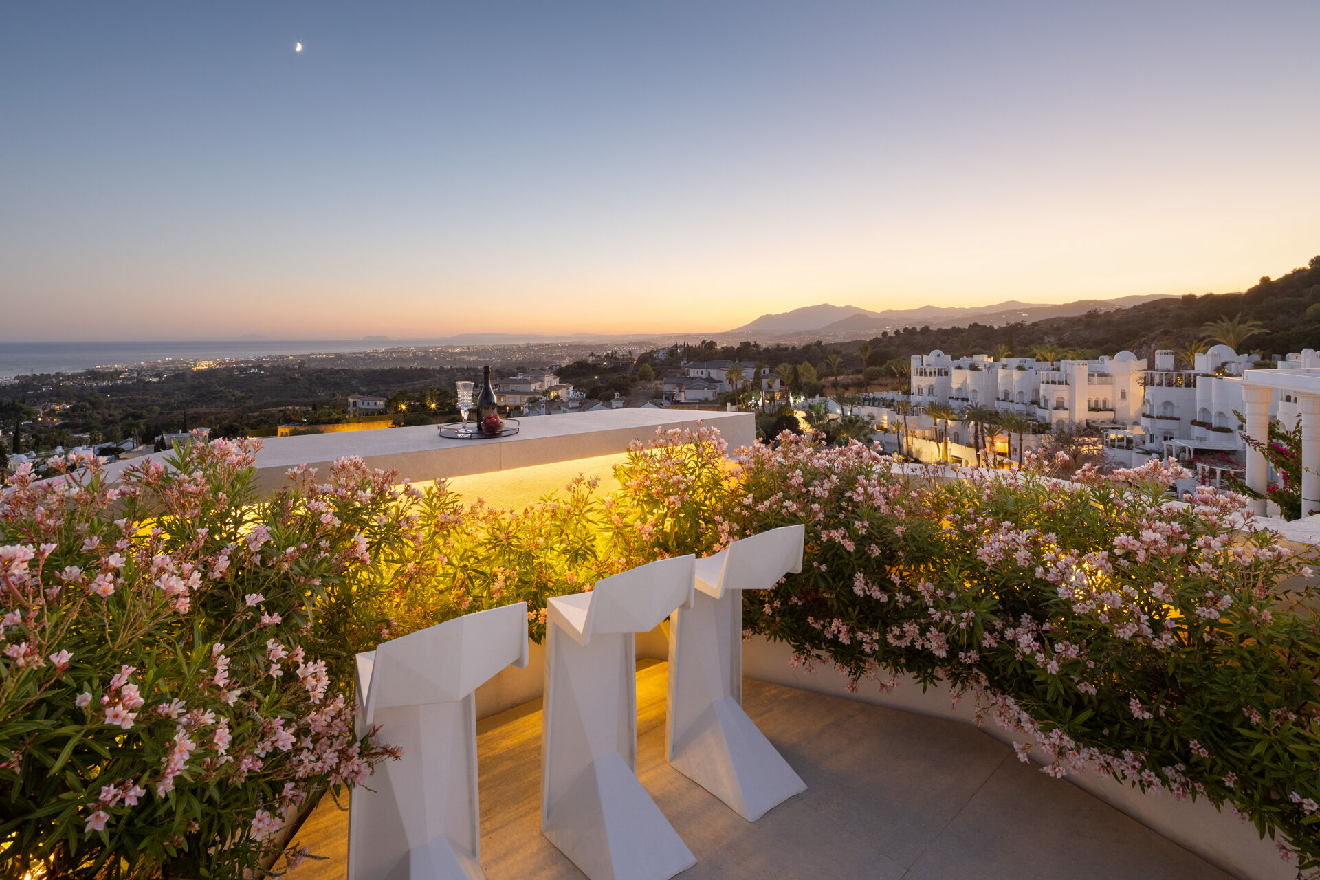 The bar counter on the roof terrace at dusk, among flowering shrubs, looking over the hillside and the coast to the sea.