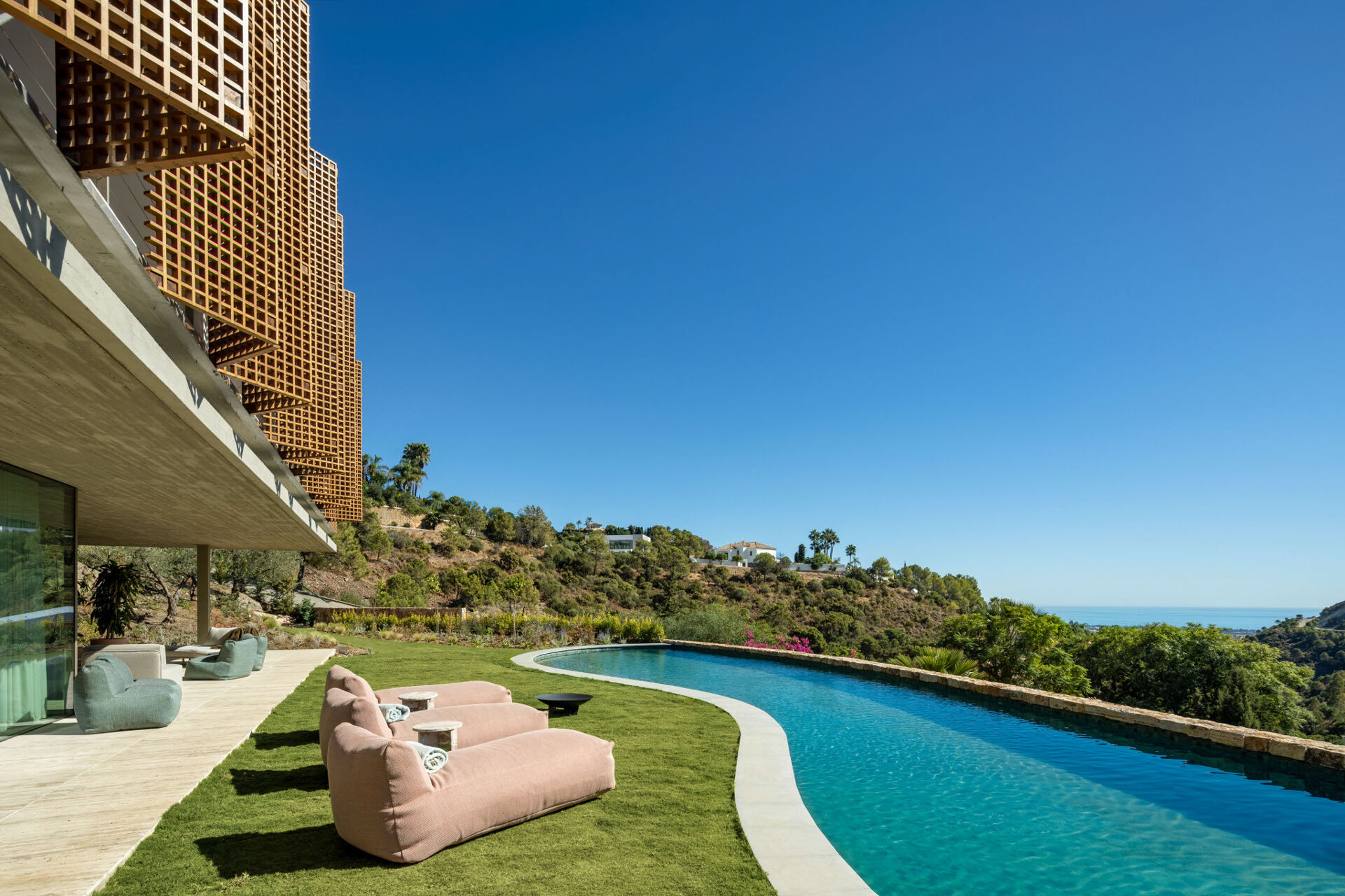 The pool curving along the edge of the lawn, with timber screens on the house above and the sea in the distance.