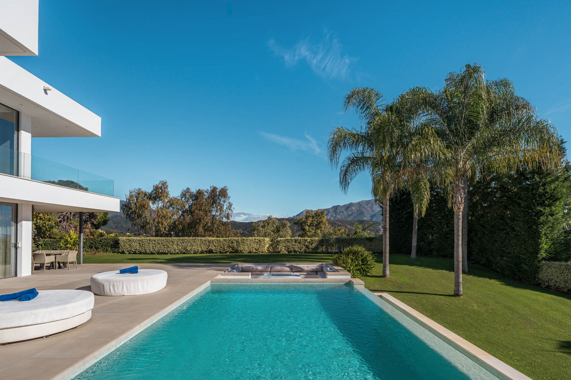 The swimming pool with a sunken seating area, the lawn and tall palms beside the house, with mountains in the distance.