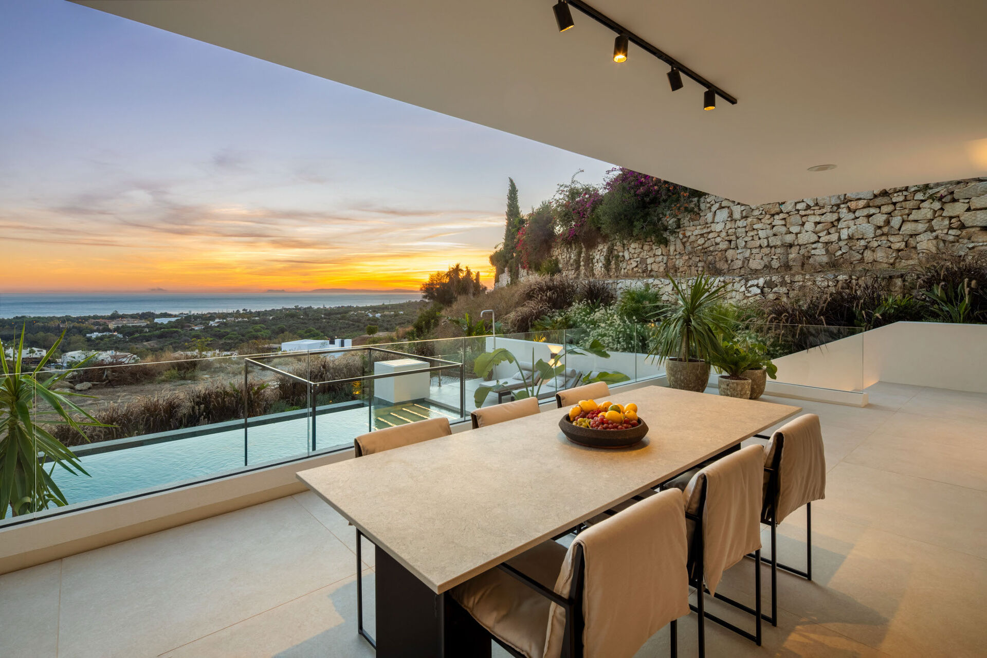 The covered terrace with a dining table, looking over the pool to the sea at sunset.