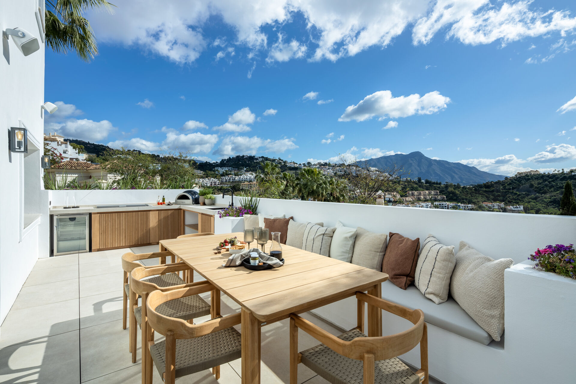 The terrace beside the pool, with an outdoor kitchen, a teak dining table and a built-in bench, and a mountain in the distance.