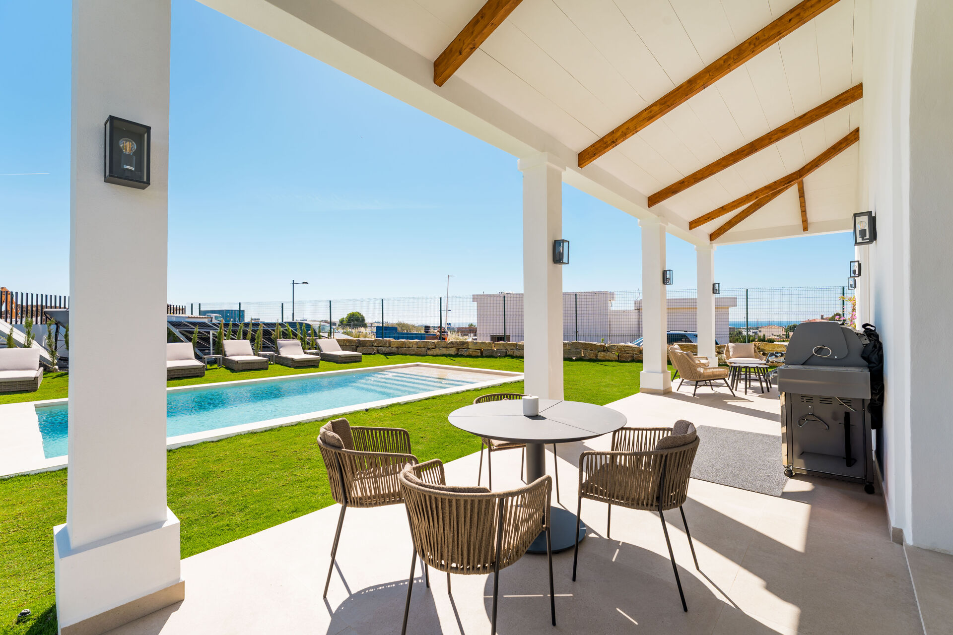 The covered terrace, with a dining table, seating and a barbecue, looking over the swimming pool and the lawn.