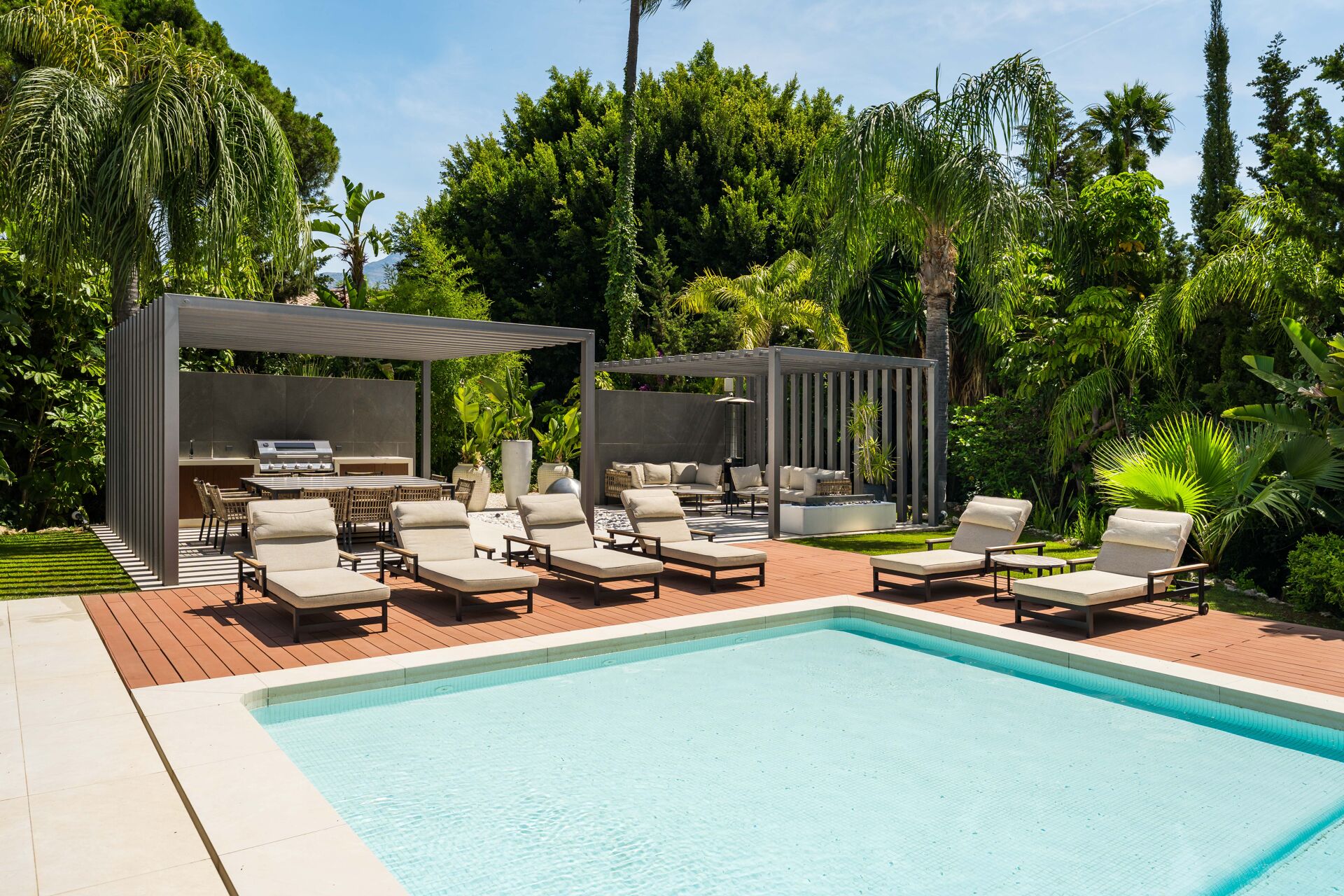 The swimming pool with sun loungers on the decking, and two pergolas in the garden, one over an outdoor kitchen and dining table and one over a sofa.