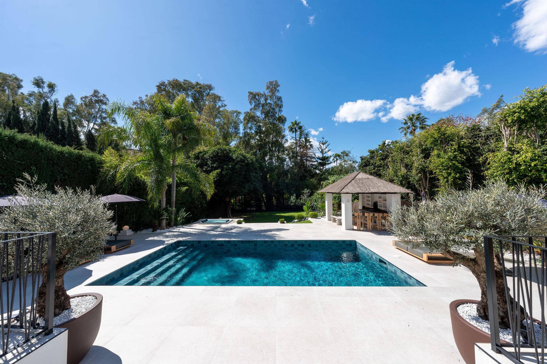 The swimming pool seen from the terrace, with the hot tub, the lawn and the outdoor kitchen pavilion beyond it.