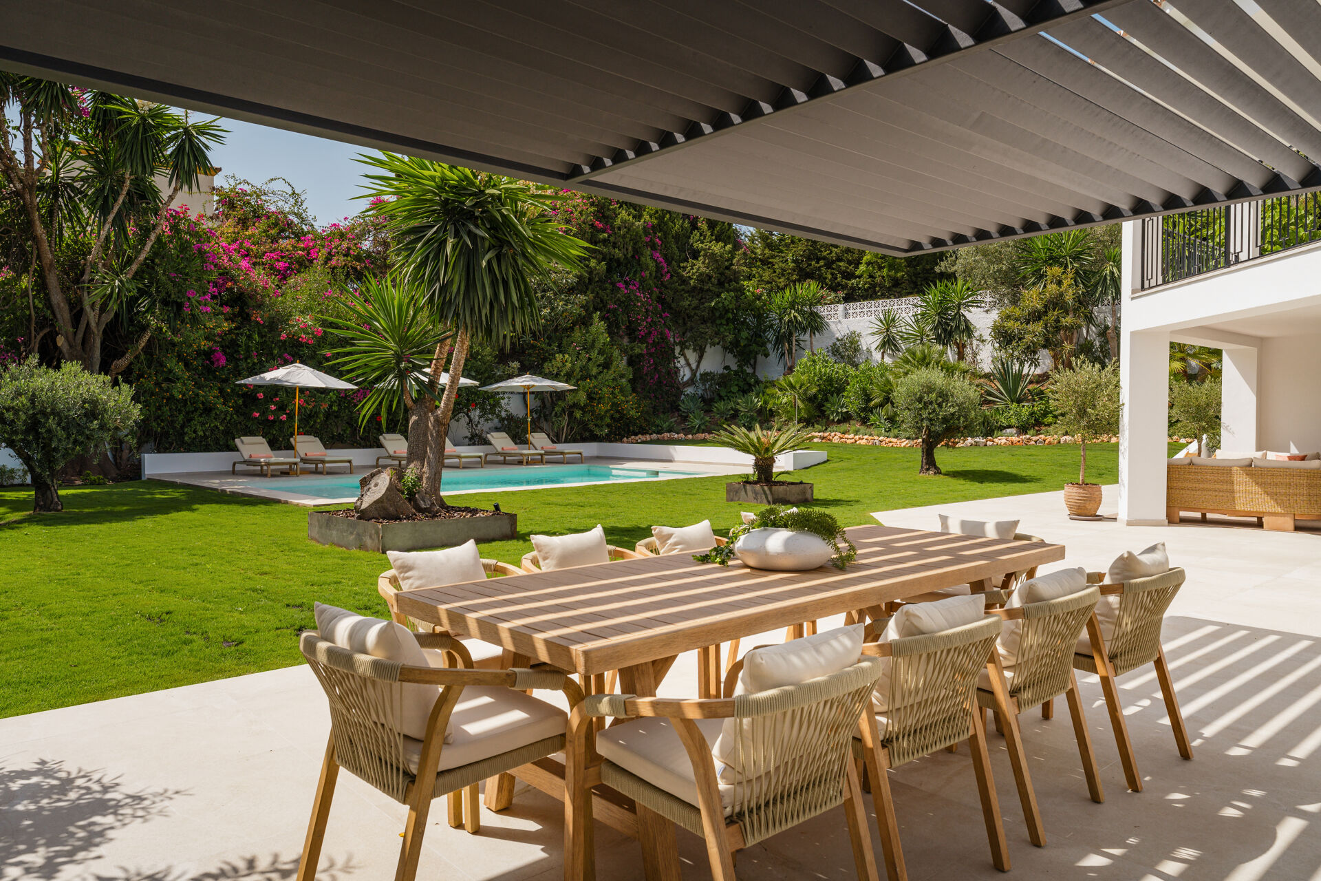 The covered dining table on the terrace, with the lawn, the swimming pool and flowering shrubs behind.